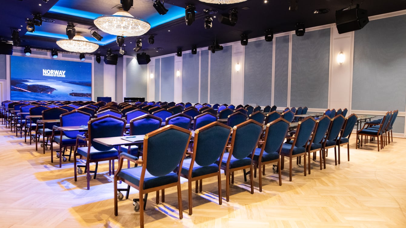 Haakon-salen i klasseromoppsett med blå stoler og store lysekroner med prosjektor og LED-skjerm i front.