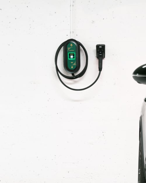 Part of a dark car in a car park with a charging cable behind it on a white wall.