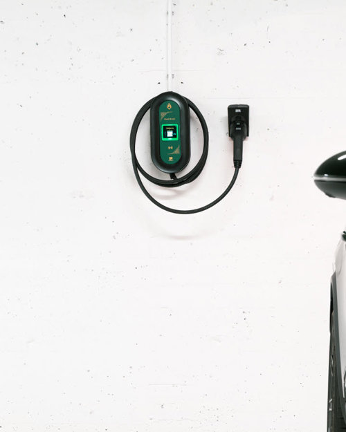 Part of a dark car in a car park with a charging cable behind it on a white wall.