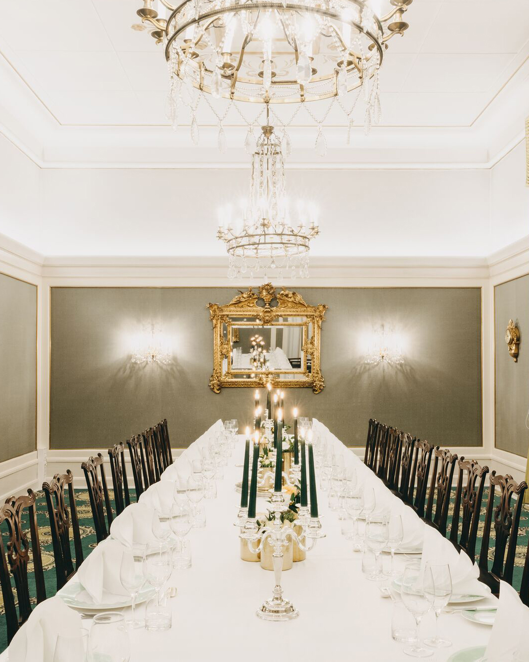 Langbord oppdekket med hvite duker, lysestaker, blomsteroppsatser. I bakgrunnen står et piano og et gullinnrammet speil. Over bordet henger to store lysekroner.