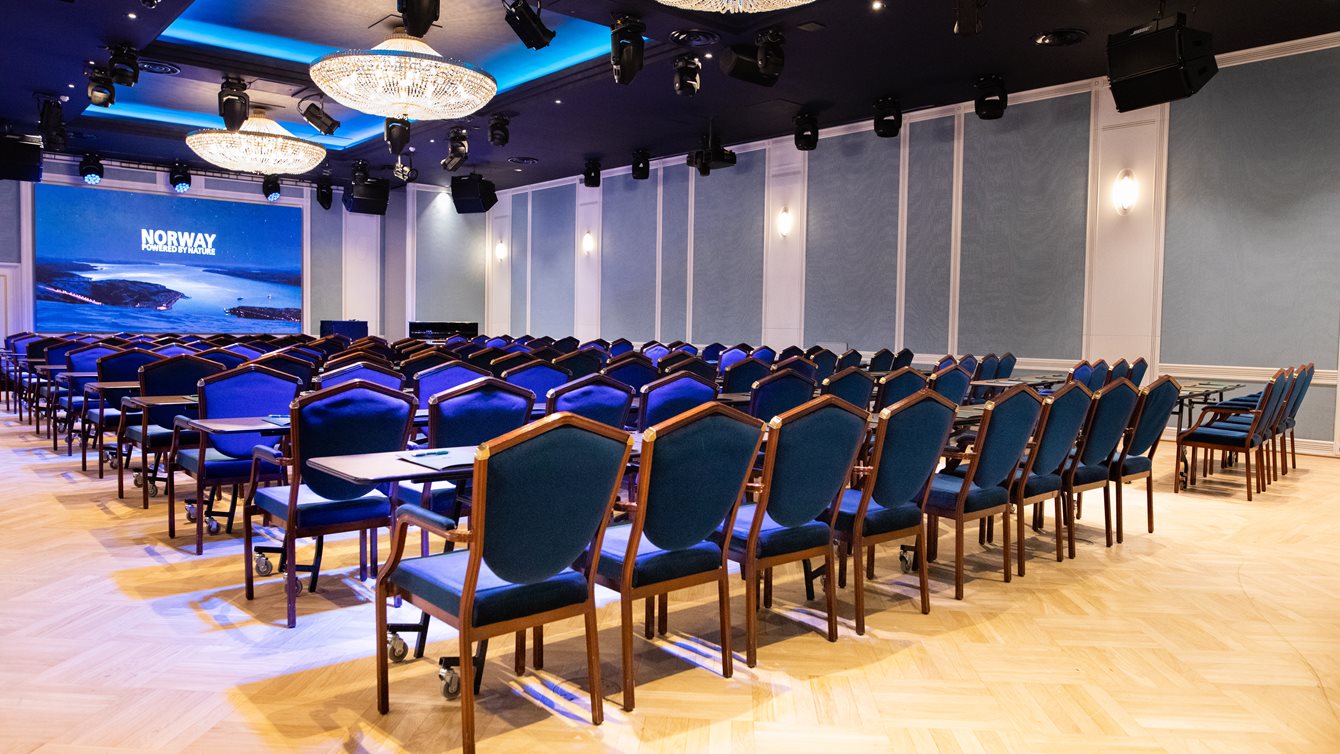 Haakon-salen i klasseromoppsett med blå stoler og store lysekroner med prosjektor og LED-skjerm i front.