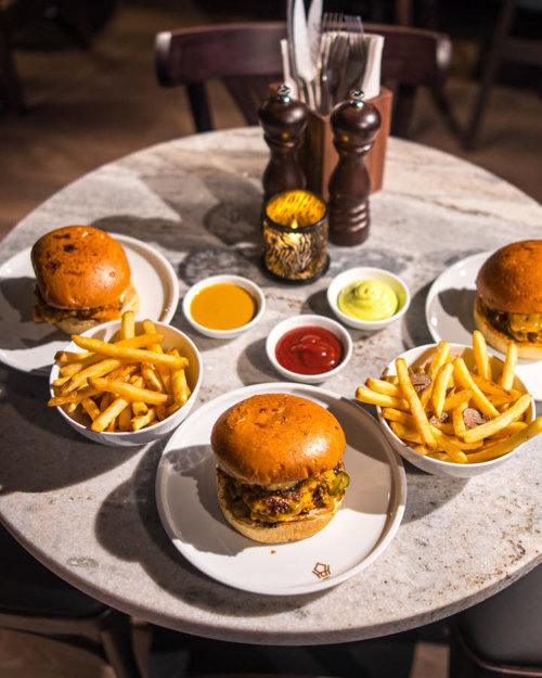 Marbmorbord med tre burgere, to skåler med pommes frites og tre ulike dipper. Salt, pepper og beholder med bestikk og servietter ses i bakgrunnen. Rundt bordet står det fire brune stoler.