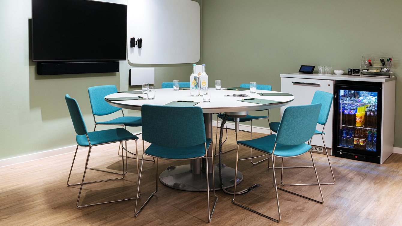 Bright meeting room with round table, turquoise chairs, paper bags and water bottle on the table. TV on the wall and whiteboard, as well as a fridge.