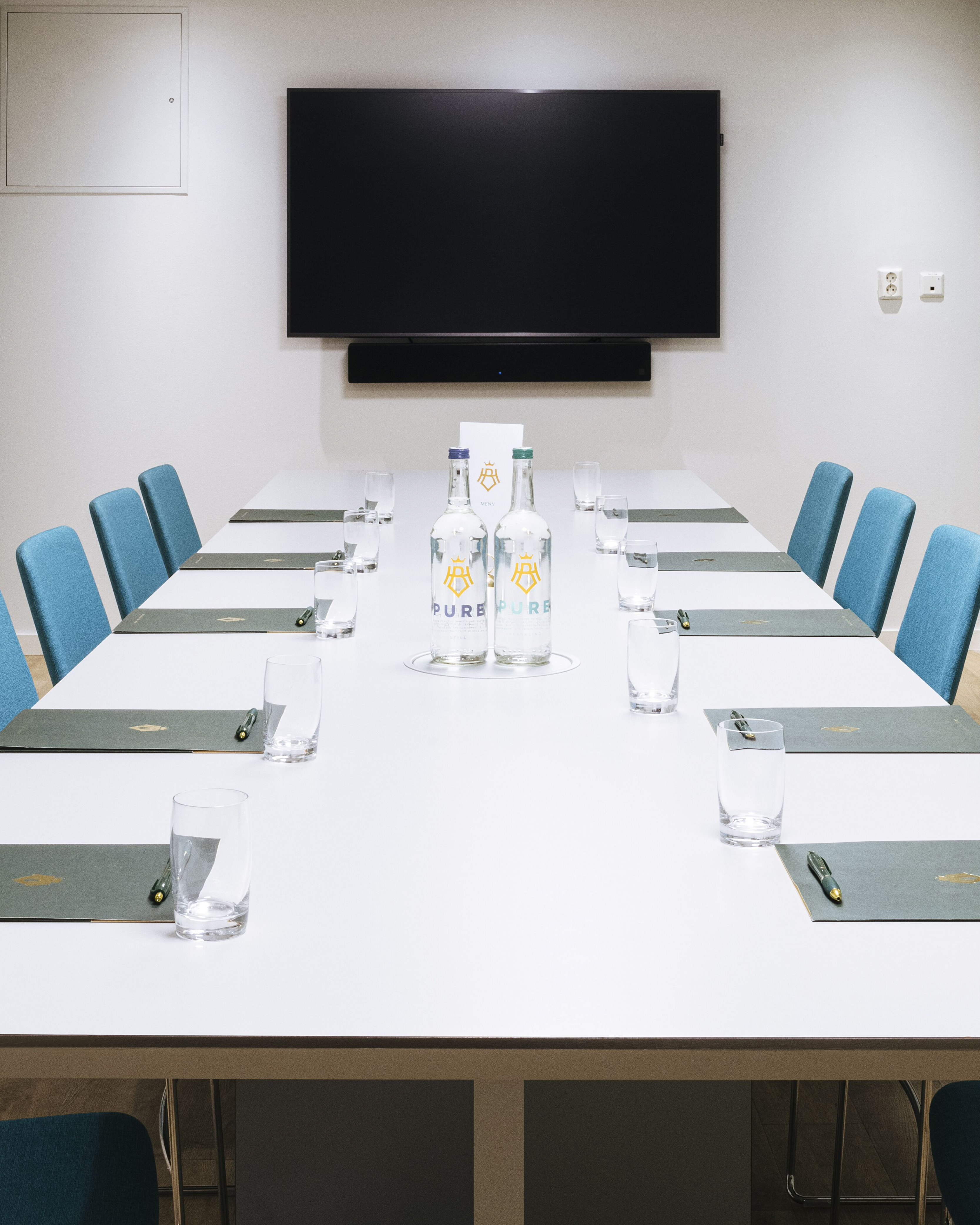 Long table with turquoise chairs, paper bags, pens, water and TV in the background