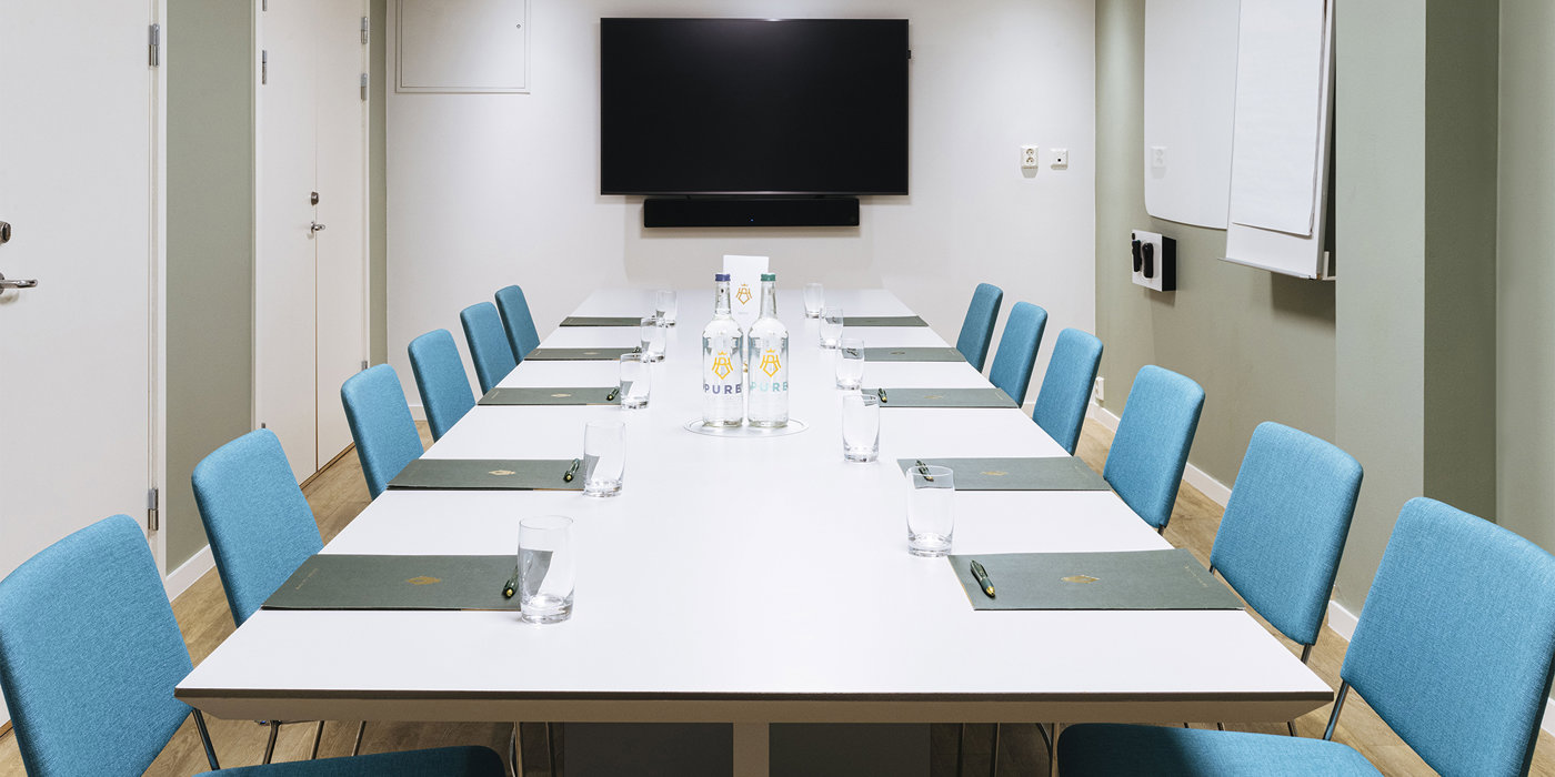 Long table with turquoise chairs, paper bags, pens, water and TV in the background