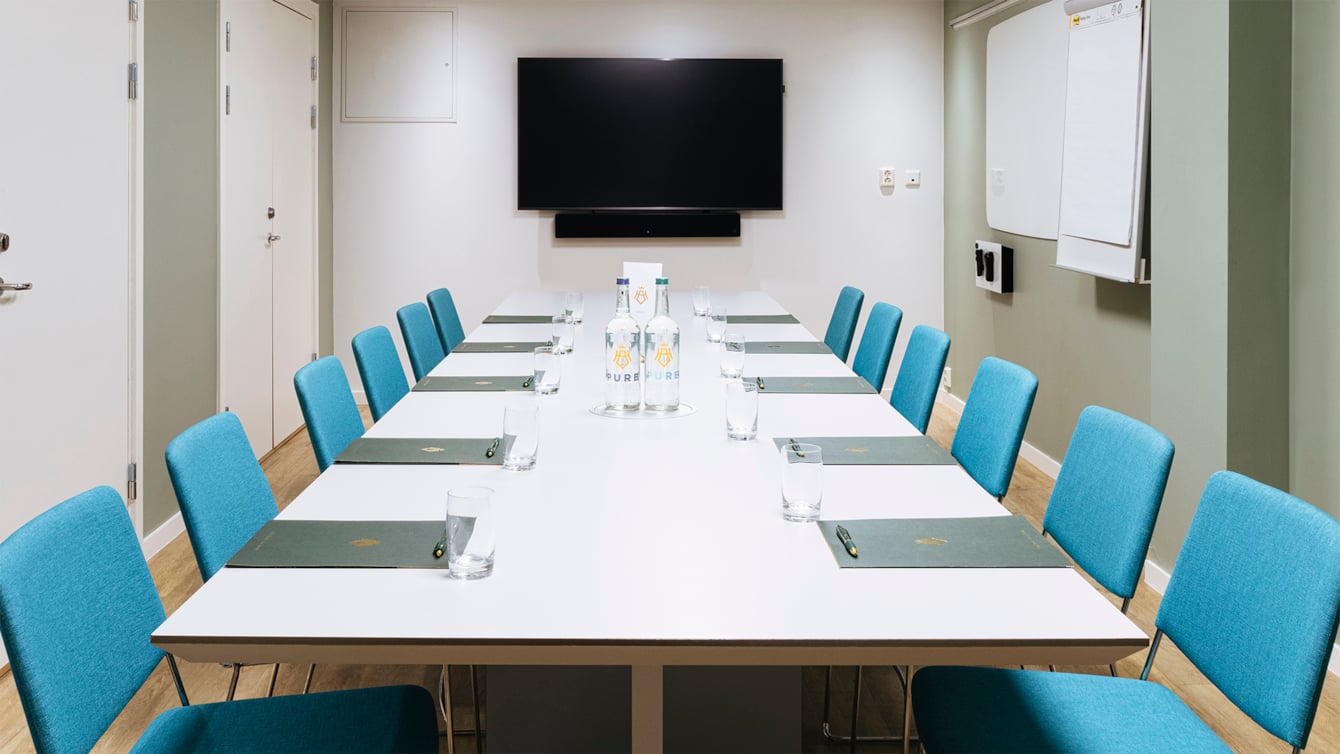 Long table with turquoise chairs, paper bags, pens, water and TV in the background