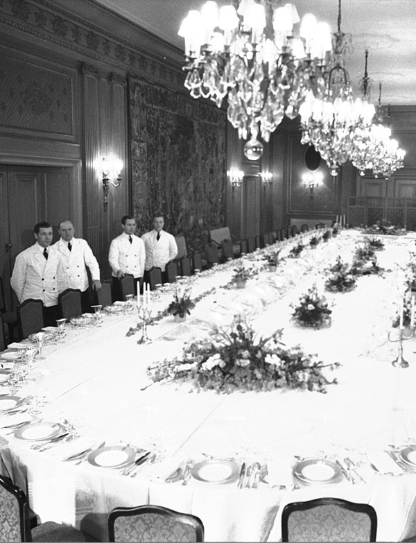 Svart-hvitt bilde av servitører i hvite uniformer på siden av et stort selskapsbord med hvite duker og oppdekket til mange gjester