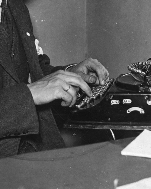 Svart-hvitt nærbilde av en hendene til en mann som skriver på en gammeldags skrivemaskin
