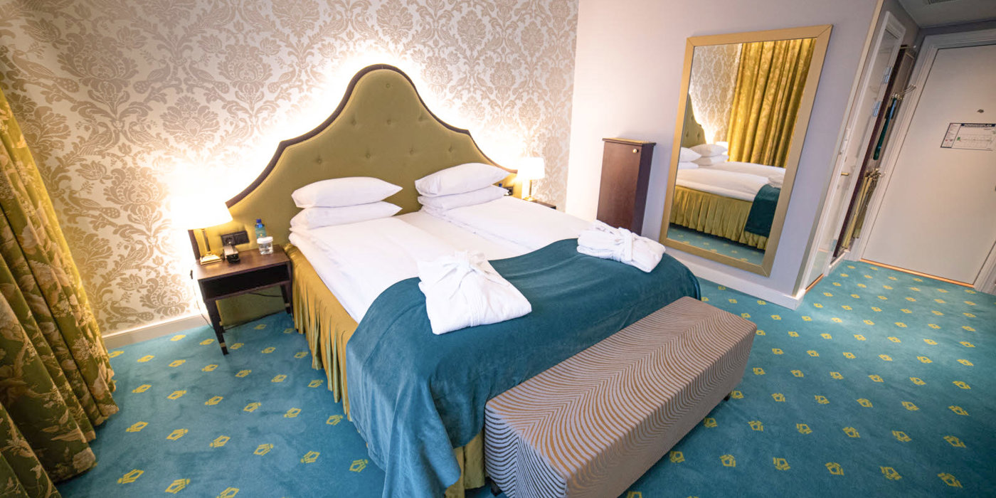 Junior suite with double bed, dressing gown, yellow headboard, patterned light wallpaper, and green wall-to-wall carpet adorned with Bristol logos.