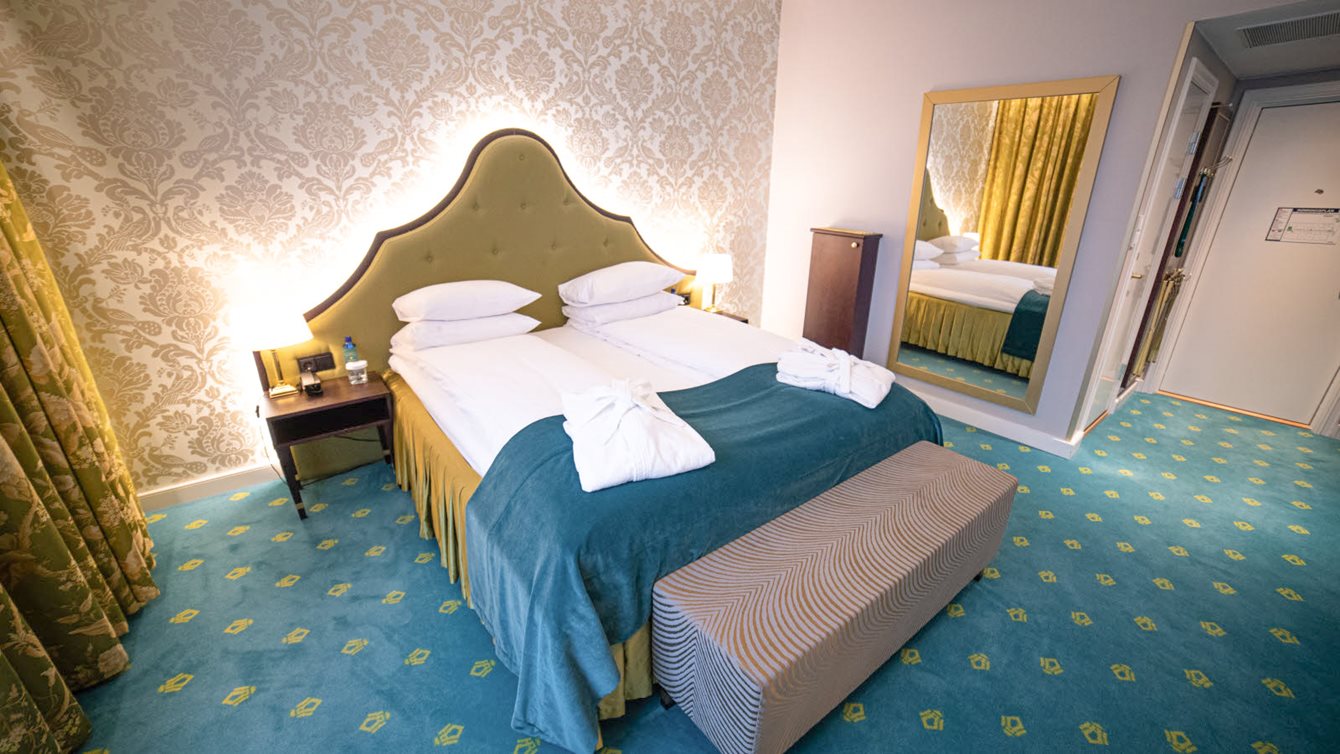 Junior suite with double bed, dressing gown, yellow headboard, patterned light wallpaper, and green wall-to-wall carpet adorned with Bristol logos.
