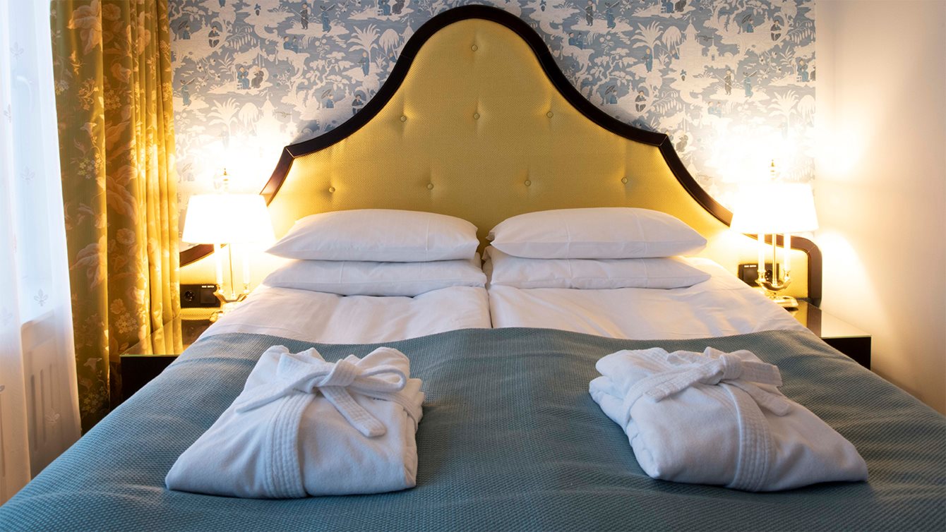King size bed with white linens, bathrobe and a golden headboard in front of a blue patterned background