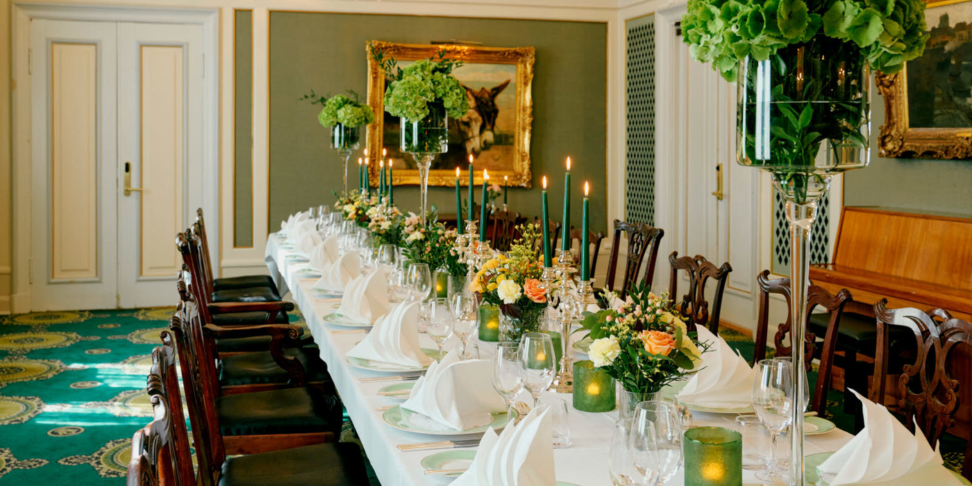 Langbord oppdekket med hvite duker, lysestaker, blomsteroppsatser. I bakgrunnen står et piano og et gullinnrammet speil. Over bordet henger to store lysekroner.
