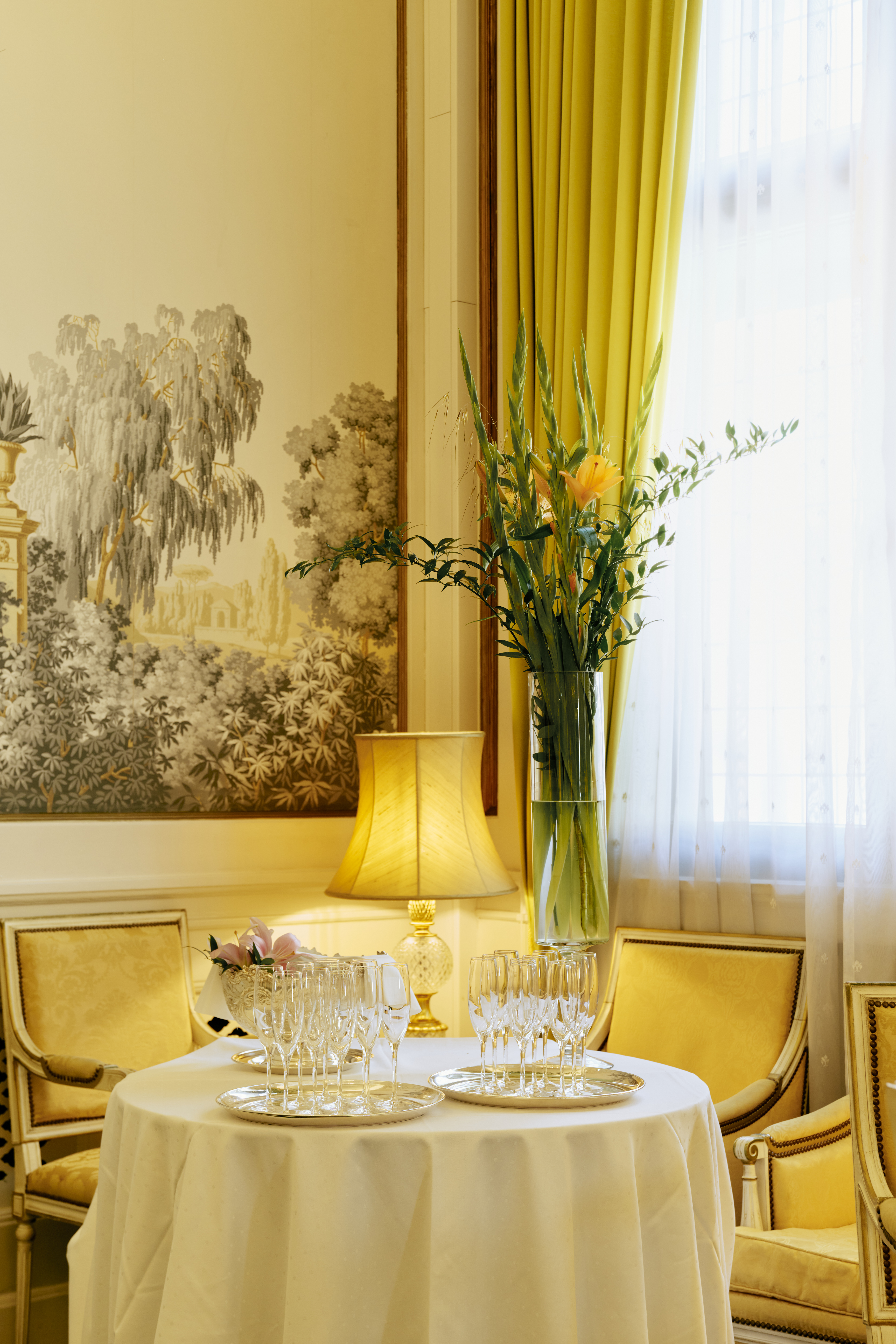 Langbord oppdekket med hvite duker, lysestaker, blomsteroppsatser. I bakgrunnen står et piano og et gullinnrammet speil. Over bordet henger to store lysekroner.