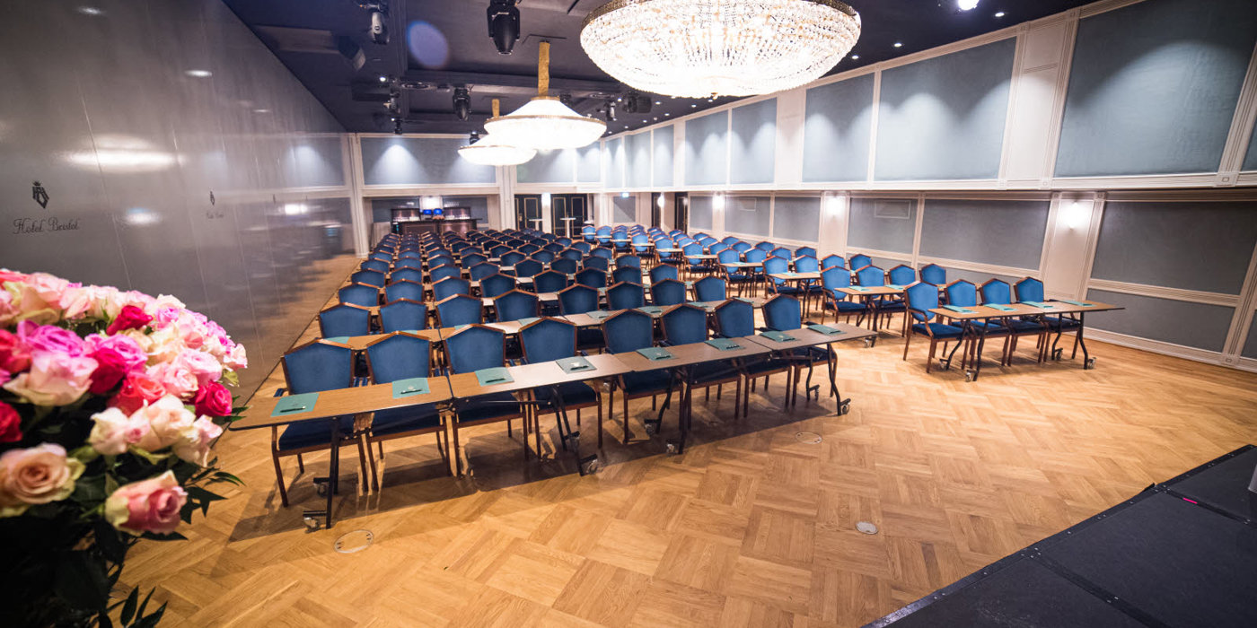 Maud-salen i klasseromoppsett med blå stoler og store lysekroner med prosjektor og LED-skjerm i front.