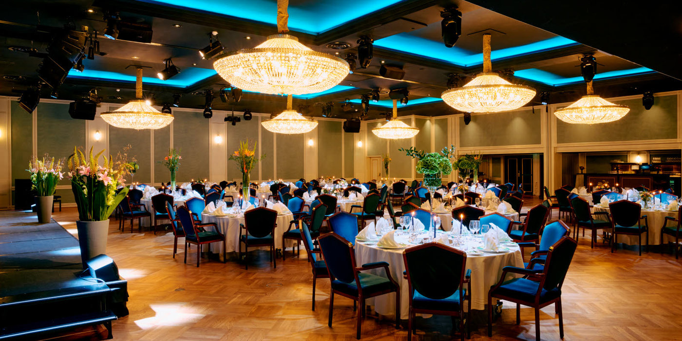 Round tables covered with white linens , two LCD screens and chandeliers hanging from the ceiling