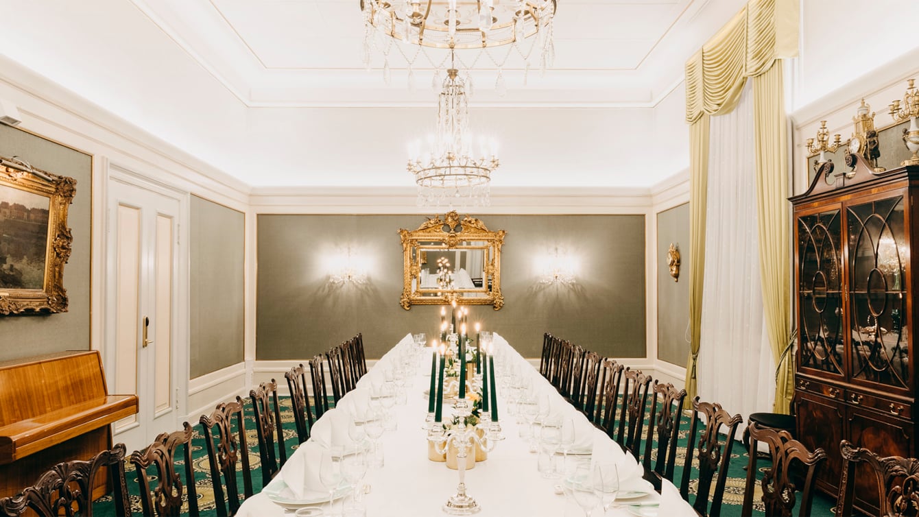 Dining table covered with white tablecloths, candlesticks, flower arrangements. In the background stands a piano and a gold-framed mirror. Two large chandeliers hanging from the ceiling