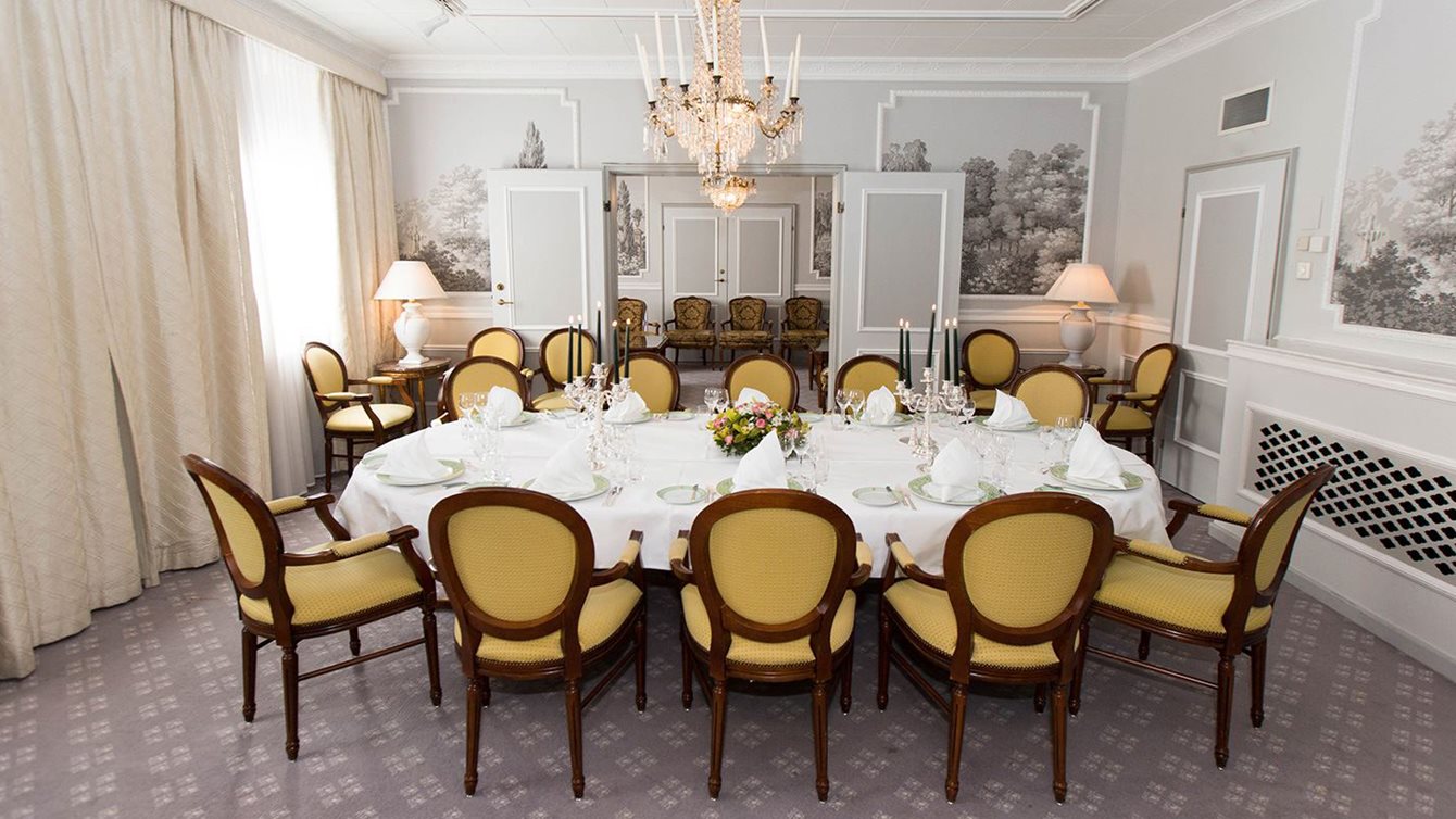 Stemningsfult og lyst selskapslokale med gule stoler, rundt langbord med hvite duker oppdekket til selskap. Over bordet henger en lysekrone