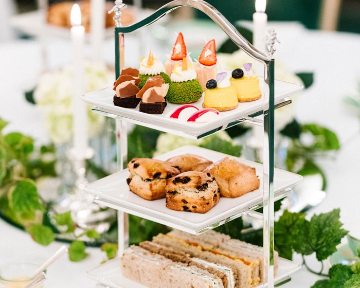 Afternoon Tea served on a triple-decker tray; mini-sandwiches on the bottom, scones in the middle and cakes on top. Next to this stand two glasses of champagne, as well as cream and jam in jars.