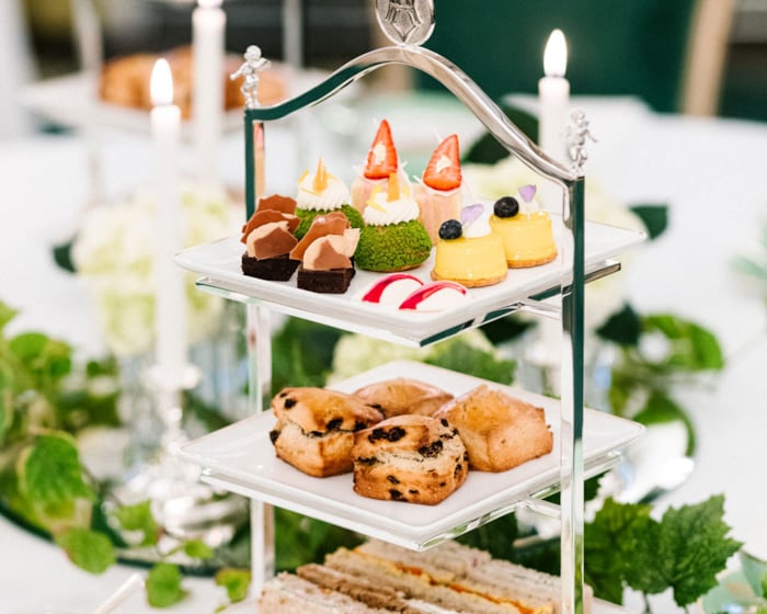 Afternoon Tea served on a triple-decker tray; mini-sandwiches on the bottom, scones in the middle and cakes on top. Next to this stand two glasses of champagne, as well as cream and jam in jars.