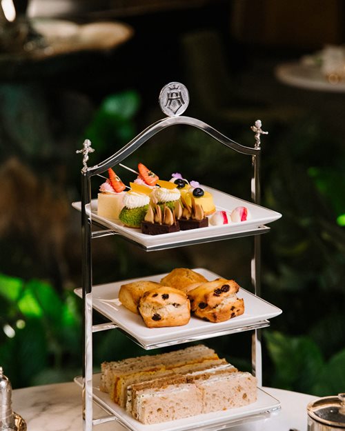 Afternoon Tea served on a triple-decker tray; mini-sandwiches on the bottom, scones in the middle and cakes on top. Next to this stand two glasses of champagne, as well as cream and jam in jars.