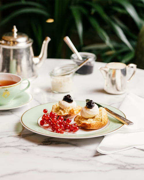 Scones toppet med luftig krem og bær med en tekanne i sølv og tekopp i bakgrunnen