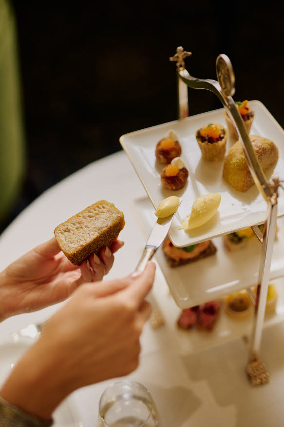 To hender som holder brødvare og kniv med smør. I bakgrunnen ses etasjefat med fingermat.