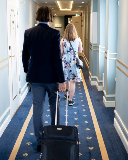 Merete og John Marius i gangen på vei til rommet sitt. Veggene i gangen er lyseblå med to horisontale gullstriper som viser vei gjennom gangen. Gulvet er teppebelagt med en mørkeblått teppe med hotellets logo i gull som rammes inn av gullstriper.
